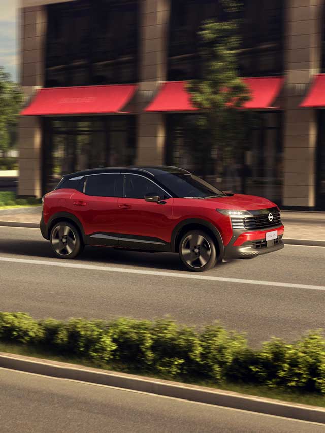 Um Nissan Kicks vermelho com teto preto em movimento em uma rua urbana. O carro é visto de perfil, passando por um prédio moderno com toldos vermelhos e árvores borradas ao fundo, indicando velocidade. O design do veículo é proeminente com sua frente angular e rodas pretas.