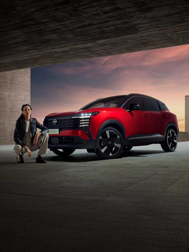 Uma pessoa se agacha ao lado de um Nissan Kicks vermelho estacionado em um espaço arquitetônico moderno e aberto ao pôr do sol, com uma iluminação dramática destacando o carro e a pessoa.
