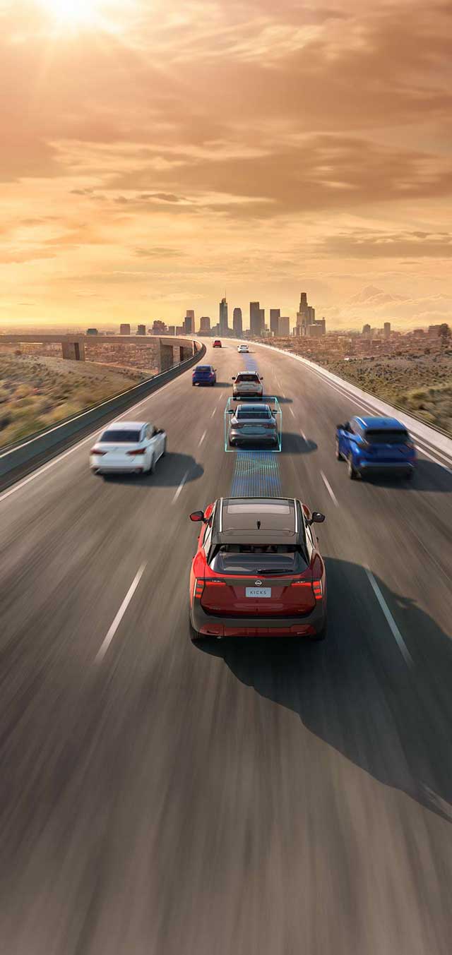 Vista aérea traseira de um Nissan Kicks vermelho em uma autoestrada urbana, com múltiplos carros em movimento, simulando tráfego. Um quadrado azul destacado mostra a ativação de um sistema de assistência à condução focando em um carro à frente. O cenário inclui uma cidade com arranha-céus ao fundo e o sol se pondo no horizonte, criando uma luz alaranjada.