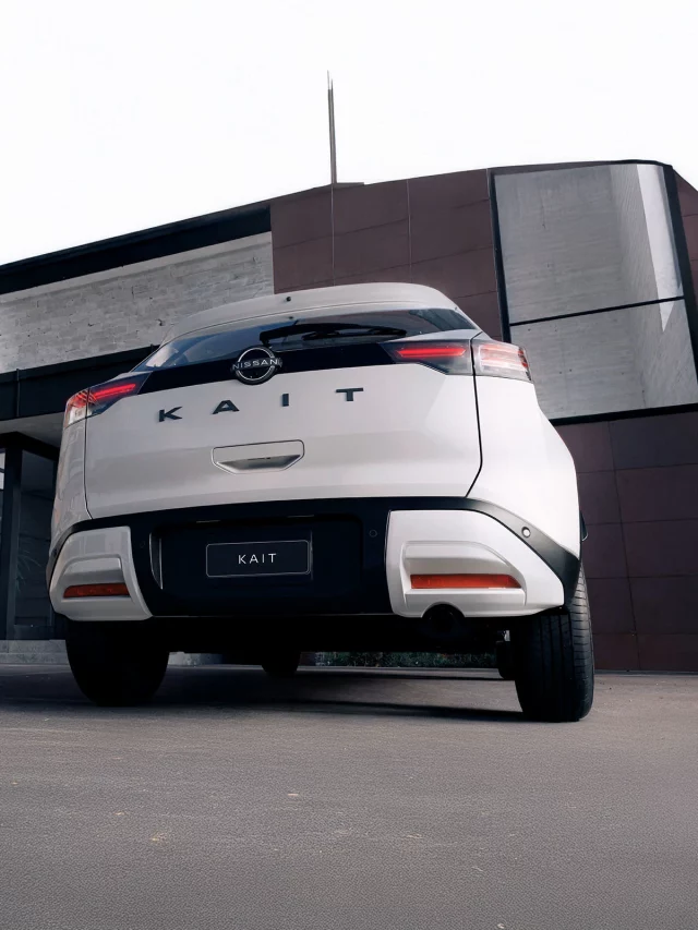 ista traseira de um Nissan KAIT branco estacionado em frente a uma residência de arquitetura moderna. As lanternas traseiras horizontais e o emblema KAIT são visíveis.