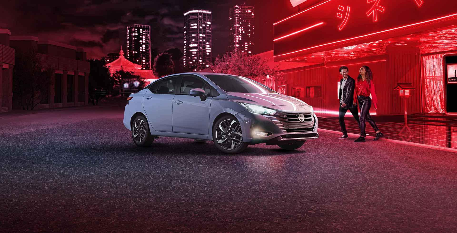 Carro Nissan Versa em uma rua iluminada por luzes vermelhas, com skyline ao fundo e duas pessoas caminhando.